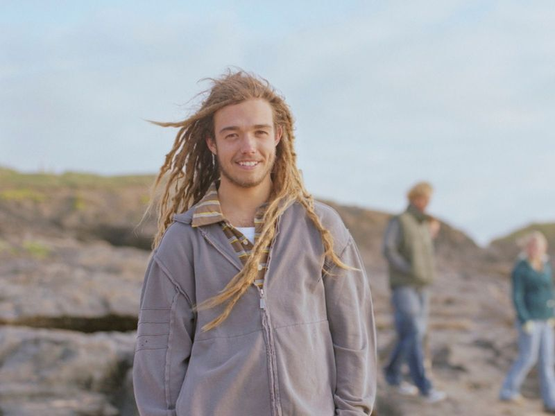 Young man standing on beach. Long hair and grey jacket