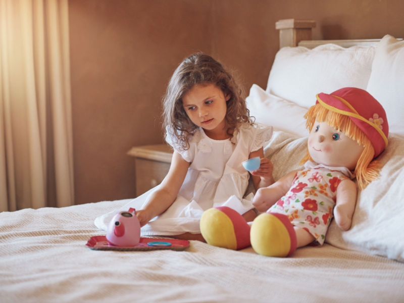 Young child playing tea sets on her bed with a large doll.