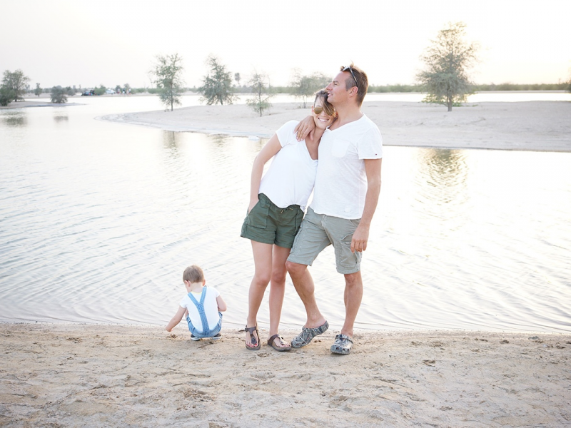 A man and woman embrace by a lake with a toddler playing at the shore