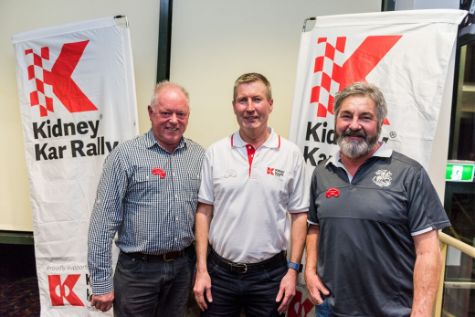 Three men pose by Kidney Kar Rally banners