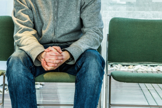 Man in green jumper, sitting in a waiting room with hands folded together.