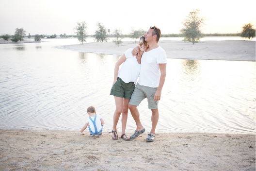 A man and woman embrace by a lake with a toddler playing at the shore